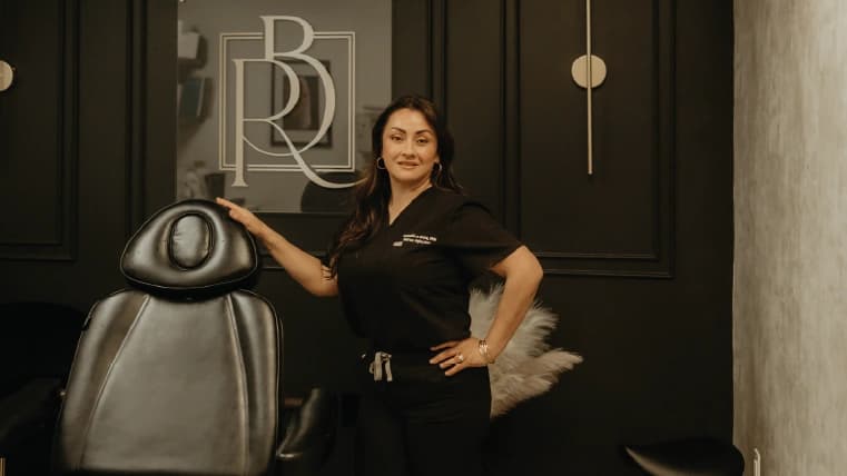 Angelica Alcaraz, BSN, RN, junto a la silla de tratamiento en el muro del logo de Revive Beauty Bar en Salinas