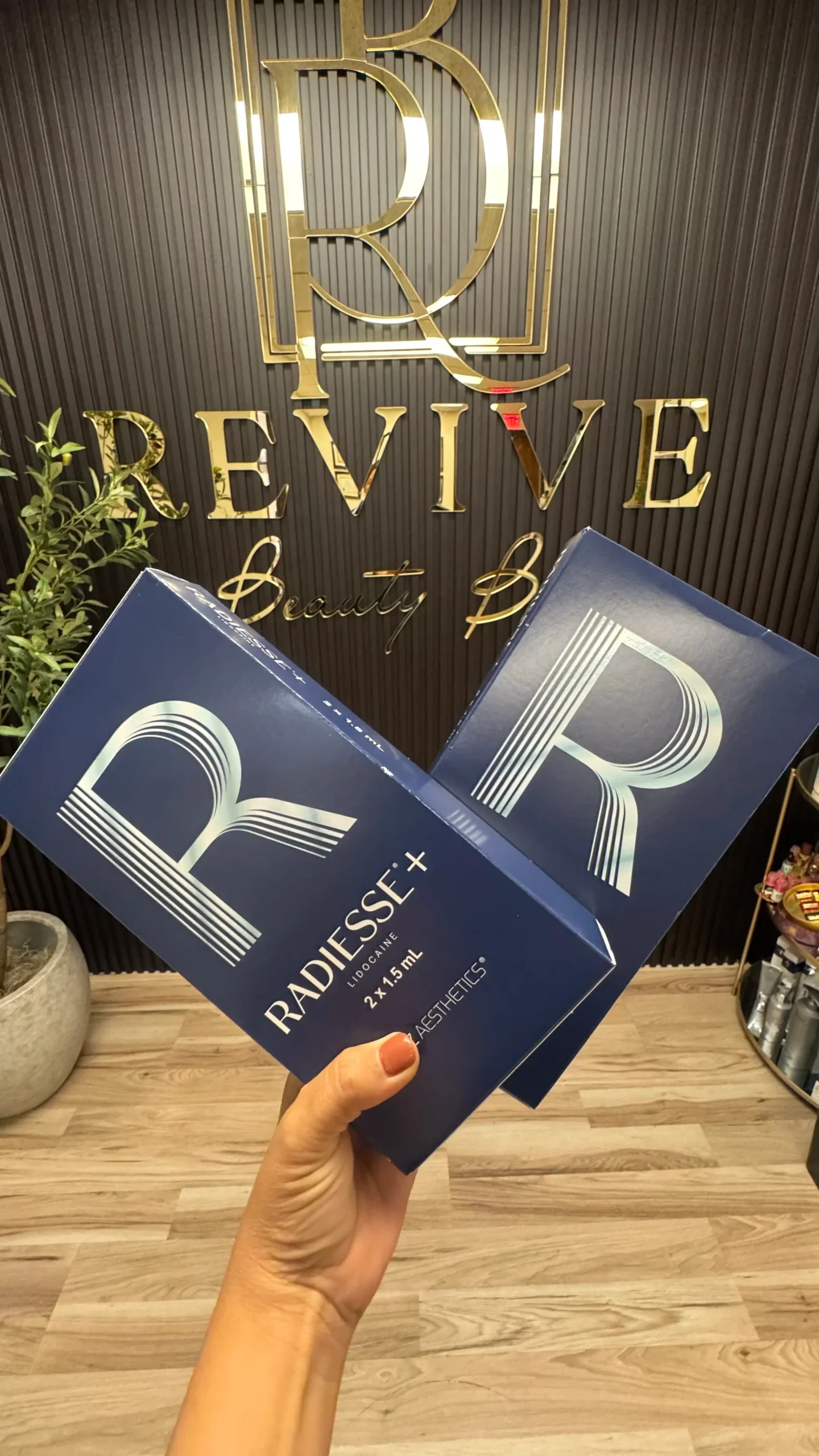 Dos cajas de Radiesse+ con lidocaína sostenidas frente al muro del logo de Revive Beauty Bar en Salinas, CA